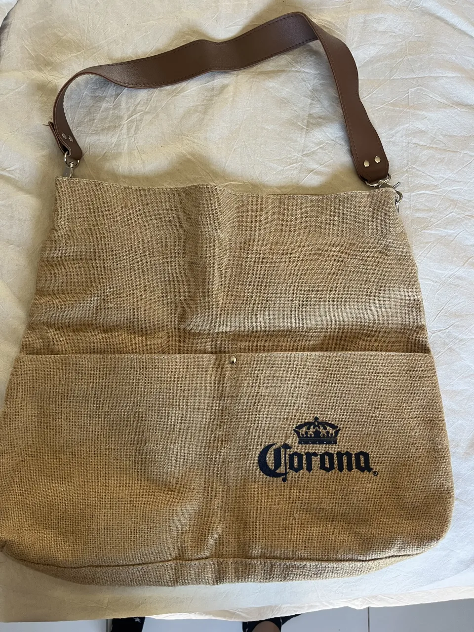 Bolso de tela de arpillera con logo de Corona estampado. Tiene una correa de cuero marrón y un bolsillo exterior.