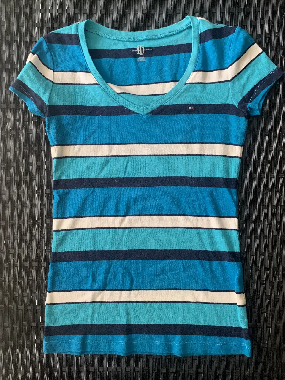 Remera Tommy Hilfiger a rayas horizontales en tonos de azul y blanco, con cuello en V y el logo de la marca bordado. Ideal para un look casual y fresco.