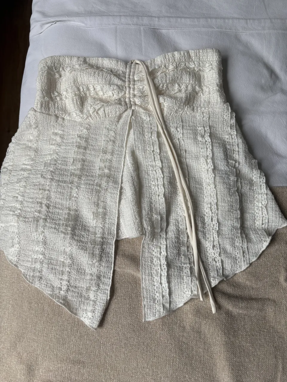 Top blanco strapless con detalle de fruncido y cordón en la parte delantera. Posee un corte asimétrico en la parte inferior.