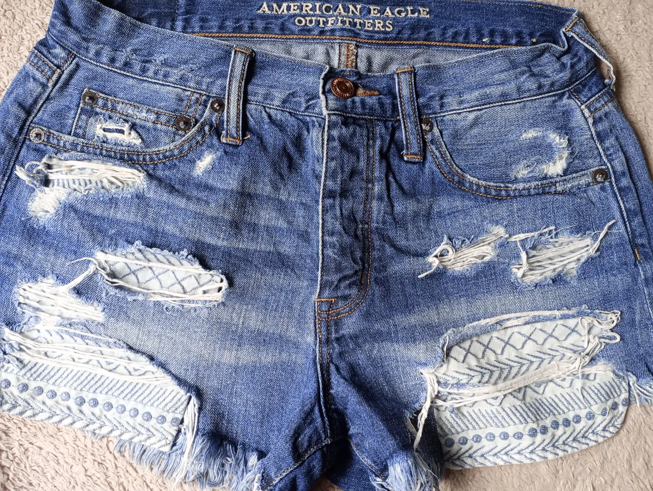 Shorts de jean tiro alto con roturas y detalles de bordado blanco en los bolsillos y en el ruedo. La parte inferior del ruedo es desflecada. talle 36