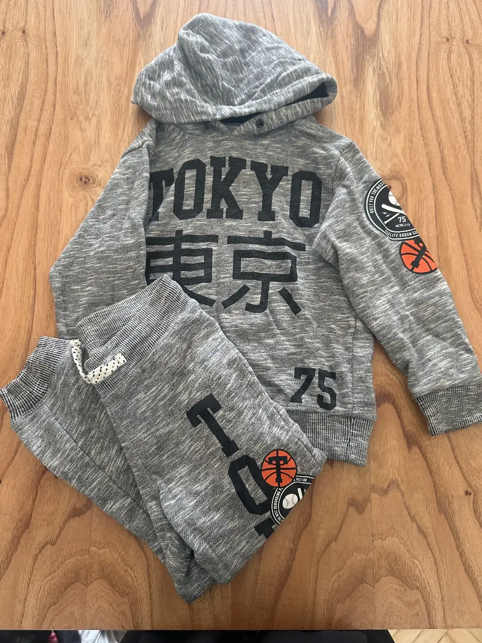 Conjunto deportivo de buzo con capucha y pantalón de jogging para nene. El buzo es de color gris con estampado "TOKYO" y detalles de baloncesto. El pantalón de jogging es gris con detalles a juego.
