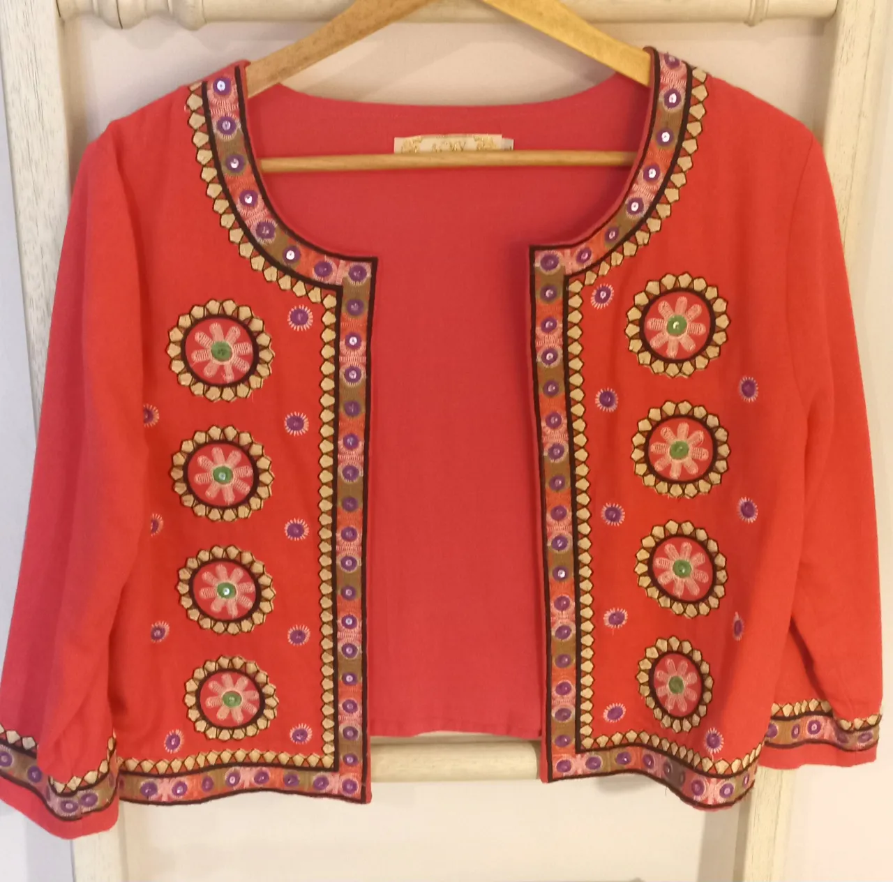 Cardigan corto de color coral con bordados florales y detalles de lentejuelas en cuello, mangas y borde inferior. Posee mangas 3/4.