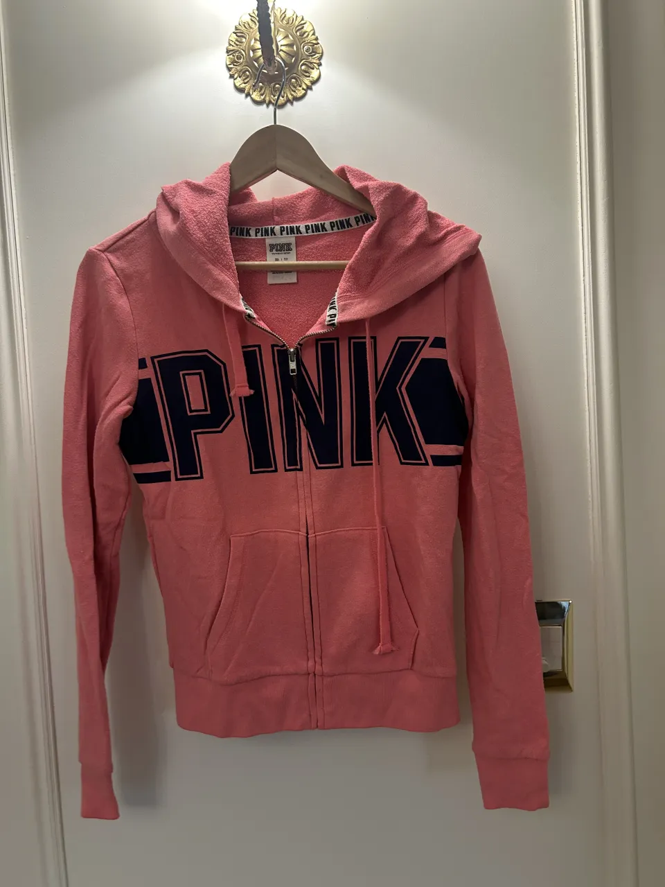 Buzo con capucha y cierre completo, de color rosa con estampa grande de letras en color azul marino en el frente. Posee bolsillos tipo canguro.