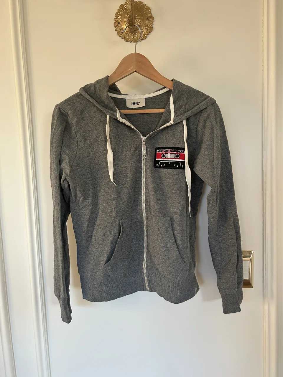 Buzo gris con capucha y cierre completo. Tiene un parche de cassette en el frente.