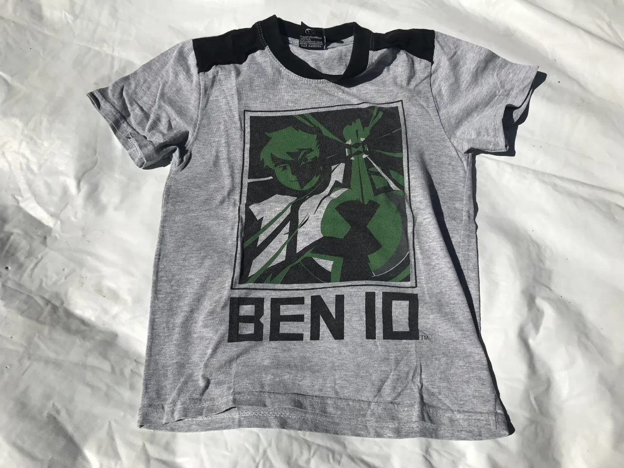 Remera gris con estampa de Ben 10 en el frente. Tiene detalles en negro en los hombros y cuello.