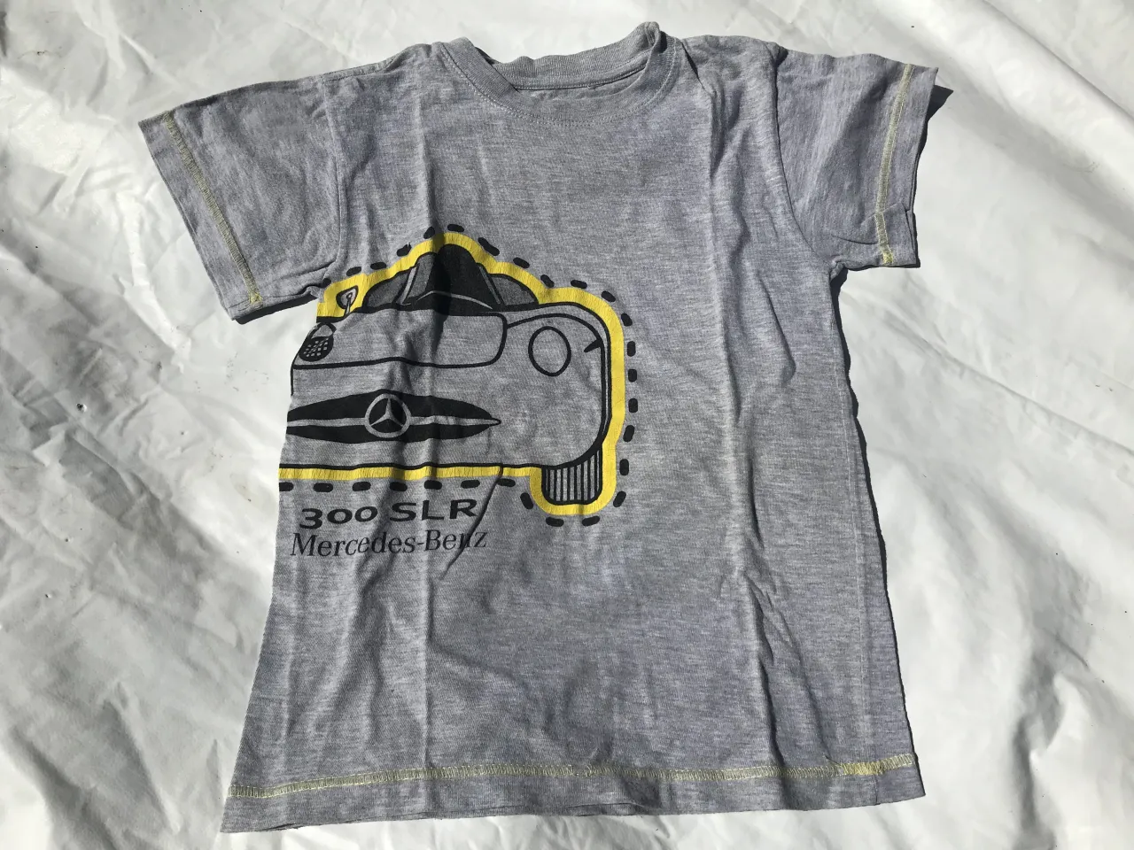 Remera gris de manga corta con estampa de un auto Mercedes-Benz 300 SLR en la parte delantera. La estampa es de color negro y amarillo.