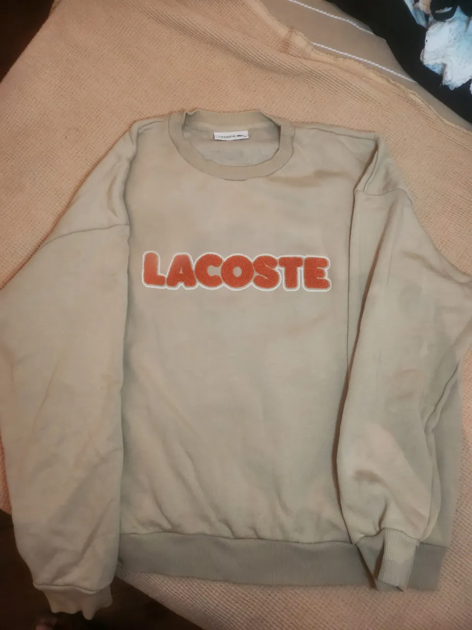 Buzo de algodón color beige con logo de Lacoste en naranja y blanco en el frente. Cuello redondo y puños elastizados.