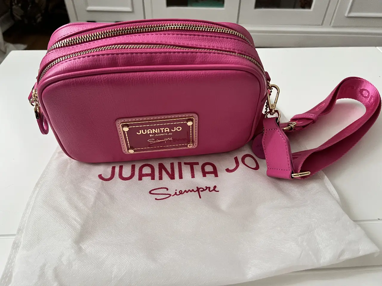 Cartera bandolera rosa fucsia de la marca Juanita Jo. Confeccionada en material sintético, presenta un diseño rectangular con doble compartimento con cierre. Detalles dorados y correa ajustable para llevar al hombro. Incluye bolsa protectora.