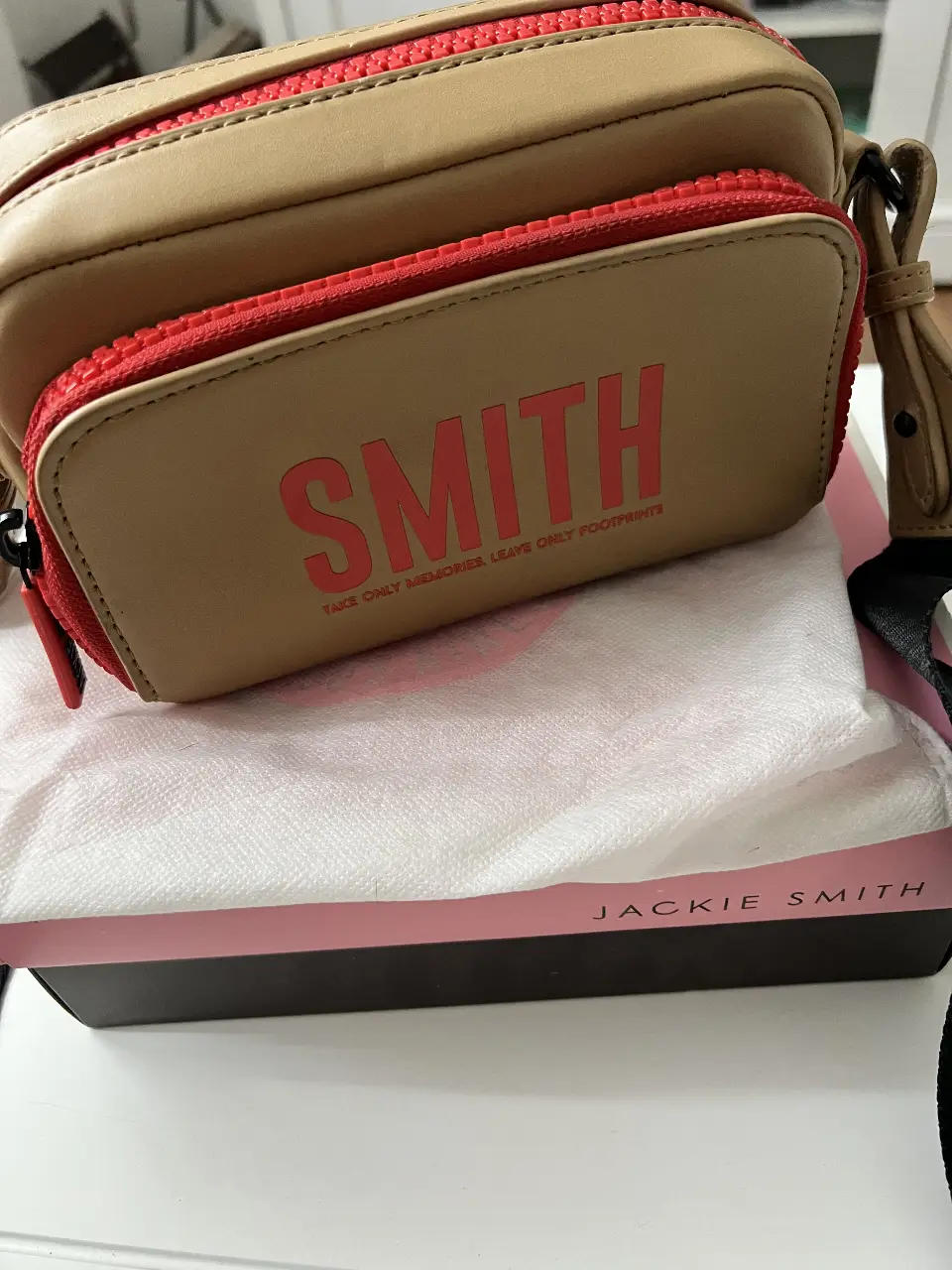 Cartera bandolera Jackie Smith, color camel con detalles en rojo. Con la inscripción SMITH en la parte frontal y cierre con detalles de la marca. Ideal para llevar tus esenciales con estilo.