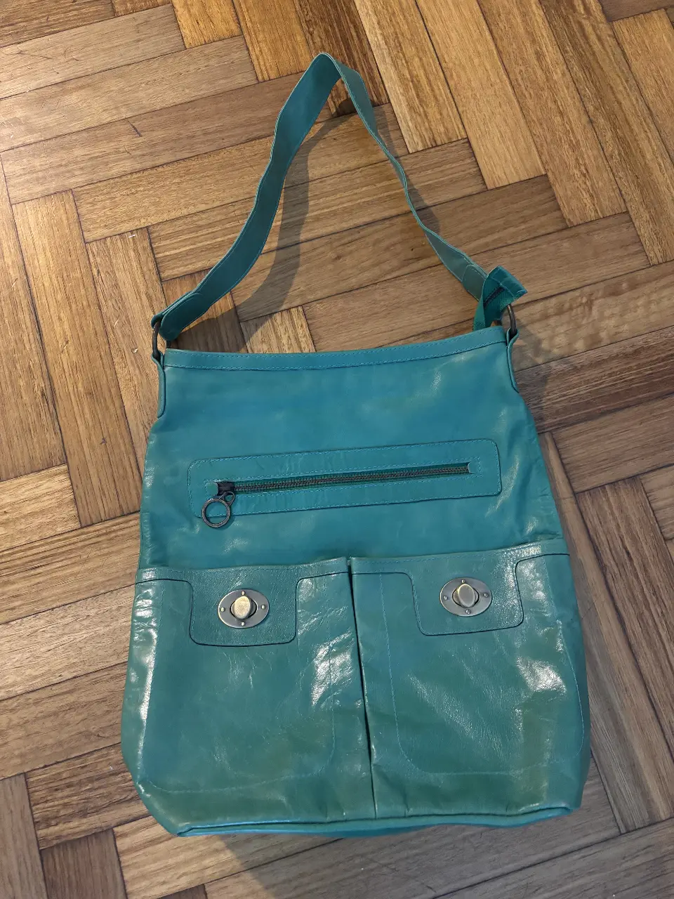 Cartera bandolera de cuero sintético color verde turquesa. Cuenta con dos bolsillos frontales con broches y un bolsillo con cierre en la parte superior. Interior con forro estampado y bolsillo interno con cierre. Ideal para un look casual y moderno.