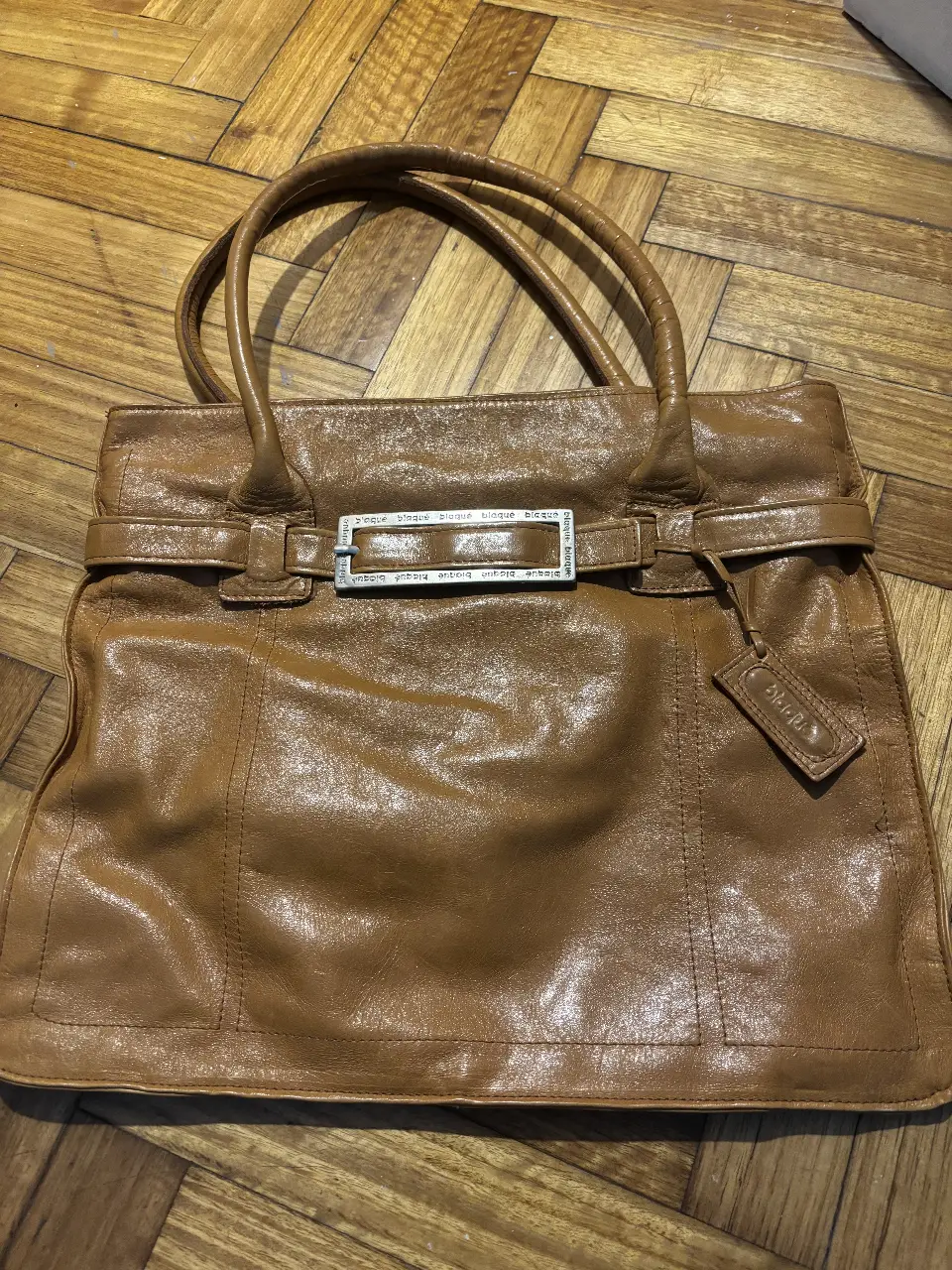 Cartera de cuero marrón de la marca Blaque. Diseño clásico con manijas cortas y correa para llevar al hombro. Detalles de hebilla y etiqueta colgante. Amplio espacio interior con forro y bolsillo interno con cierre. Ideal para uso diario.