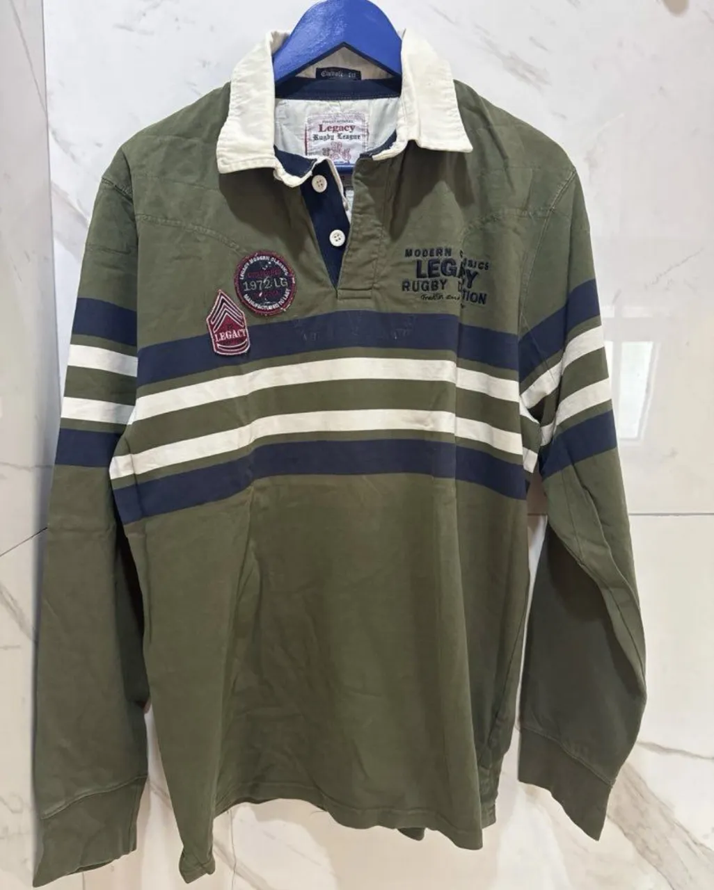 Remera manga larga de rugby, color verde militar con rayas horizontales en azul marino y blanco. Cuello blanco con detalles en azul. Estilo deportivo y casual, ideal para el día a día.