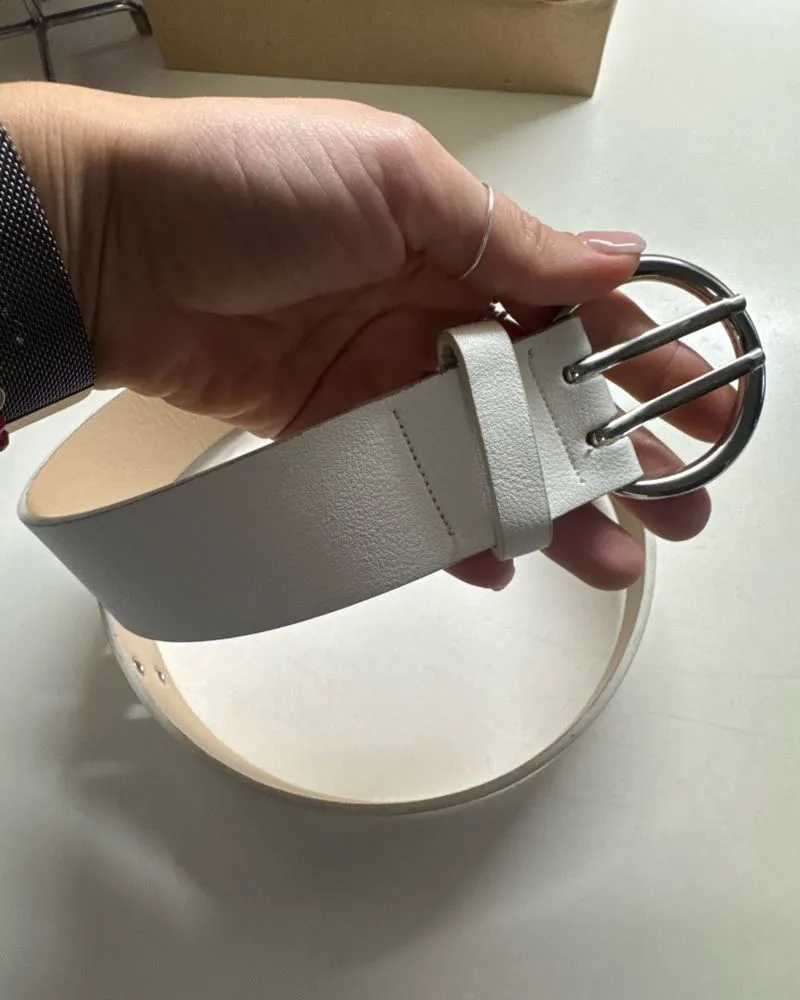 Cinturón blanco con hebilla plateada redonda y doble pasador. Posee múltiples ojales metálicos a lo largo de la tira.