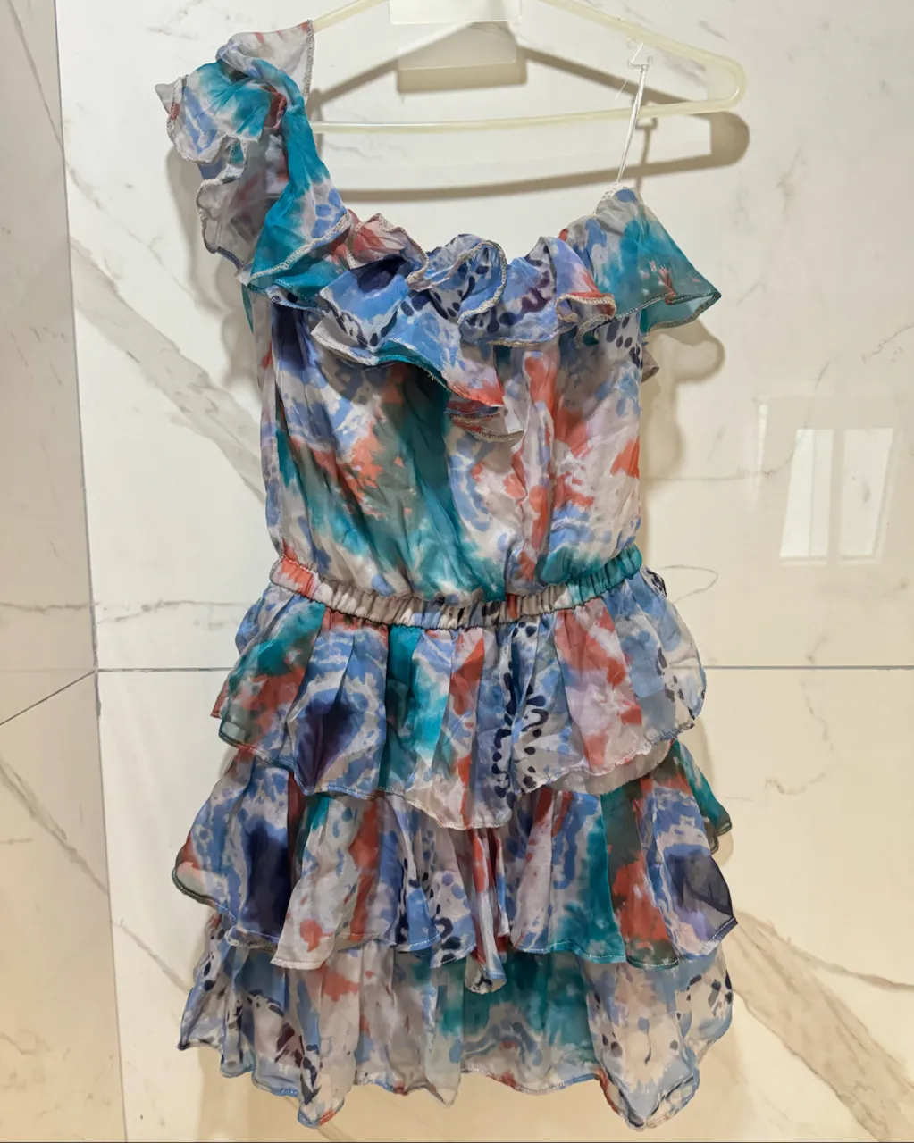 Vestido corto con volados y estampa floral abstracta en tonos azules, celestes, rosas y blancos. Diseño asimétrico con un solo hombro y volados en cascada.