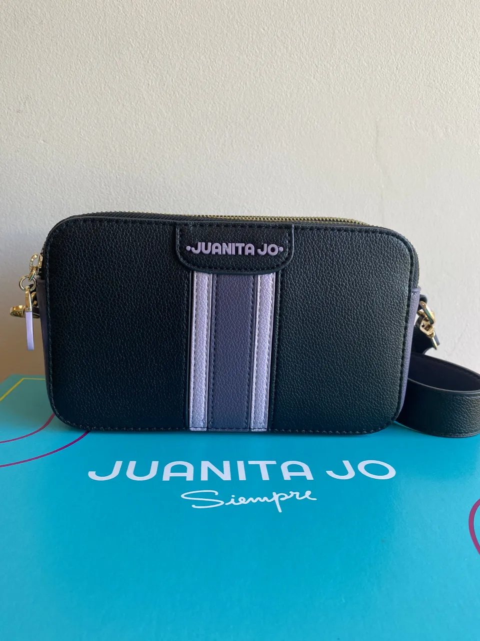 Cartera negra con detalles de tiras verticales en color lila y gris. Tiene cierre superior y correa desmontable. La marca 'JUANITA JO' se encuentra estampada en la parte frontal y en la bolsa de guardado.