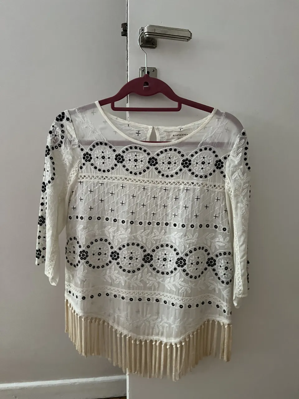 Blusa blanca con mangas 3/4 y detalles bordados en negro. Presenta un diseño con círculos y cruces, y flecos en la parte inferior.