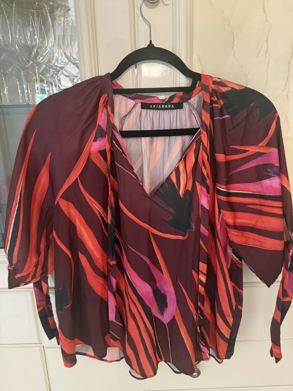 Blusa Akiabara,  con escote en V y lazo en el cuello. Posee mangas cortas con volumen, con puños atados y estampa abstracta en tonos de rojo, naranja, rosa y negro.Sin uso.