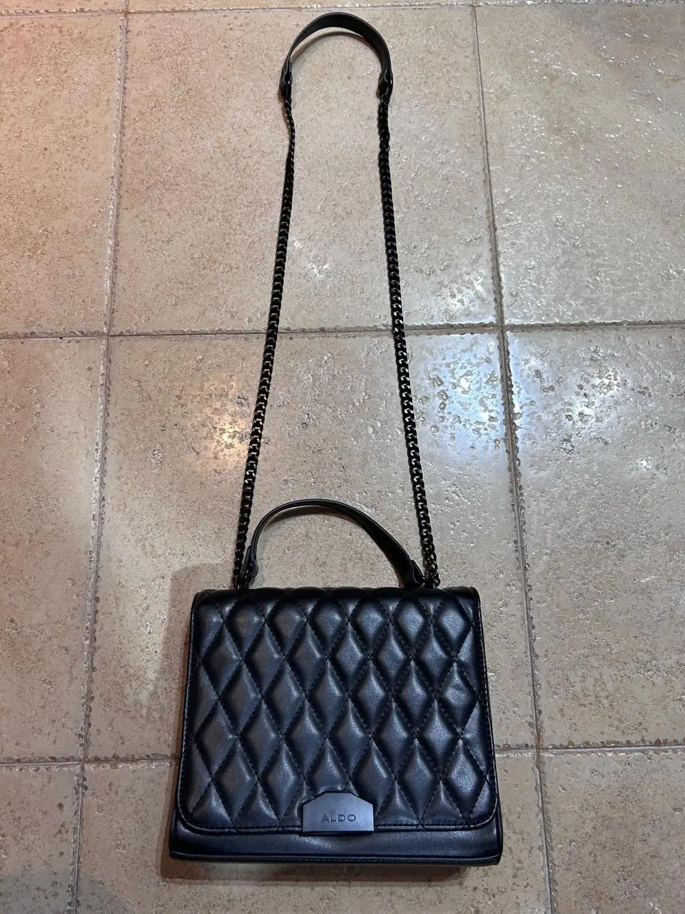 Cartera negra con diseño de rombos acolchados, con cadena para colgar y manija superior. Ideal para un look elegante y moderno.