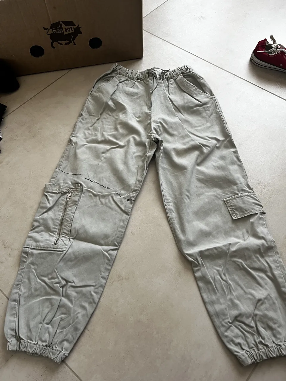 Pantalón cargo de color gris claro con bolsillos laterales y cierre en uno de ellos. Cintura elastizada con cordón. tiene en la pierna derecha una raya con marcador 