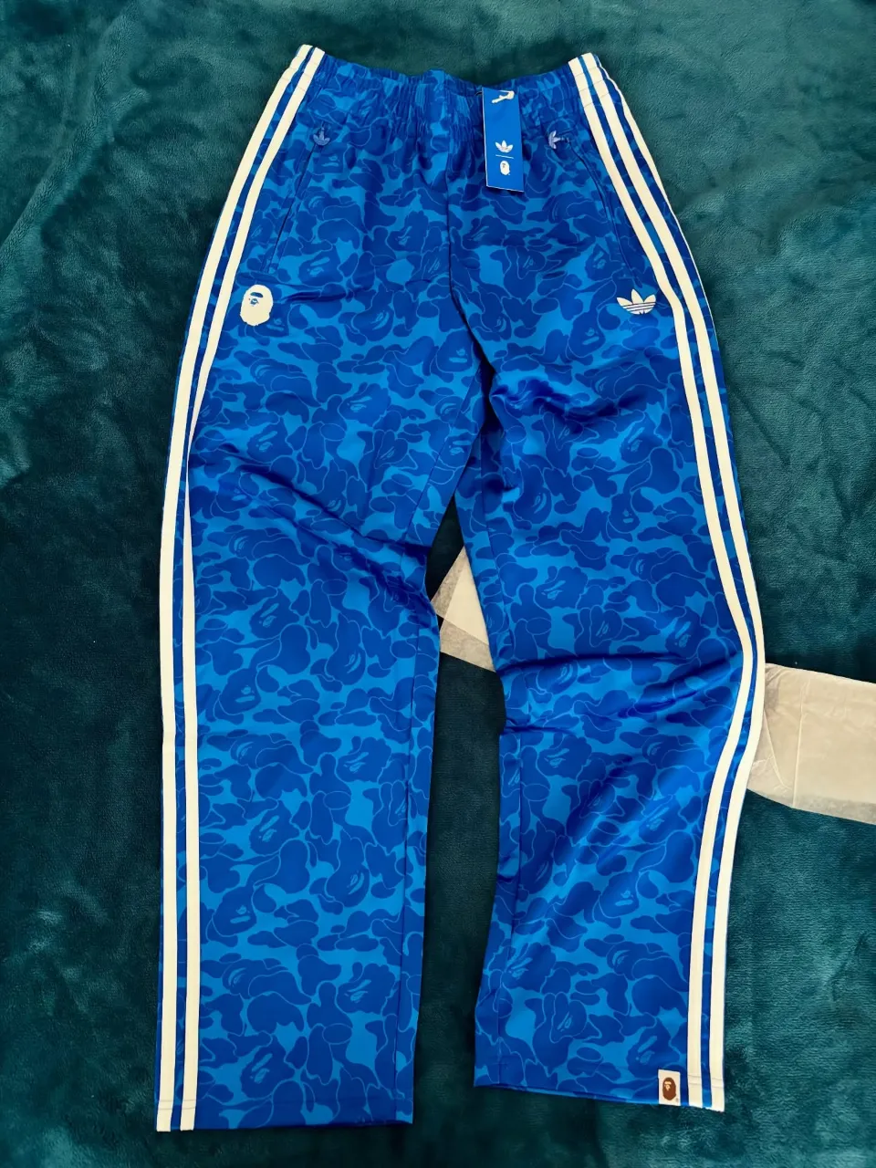 Pantalón de acetato azul con estampa camuflada en tono más claro. Posee tres tiras blancas laterales y logos de Adidas y Bape estampados. Cintura con elástico.
