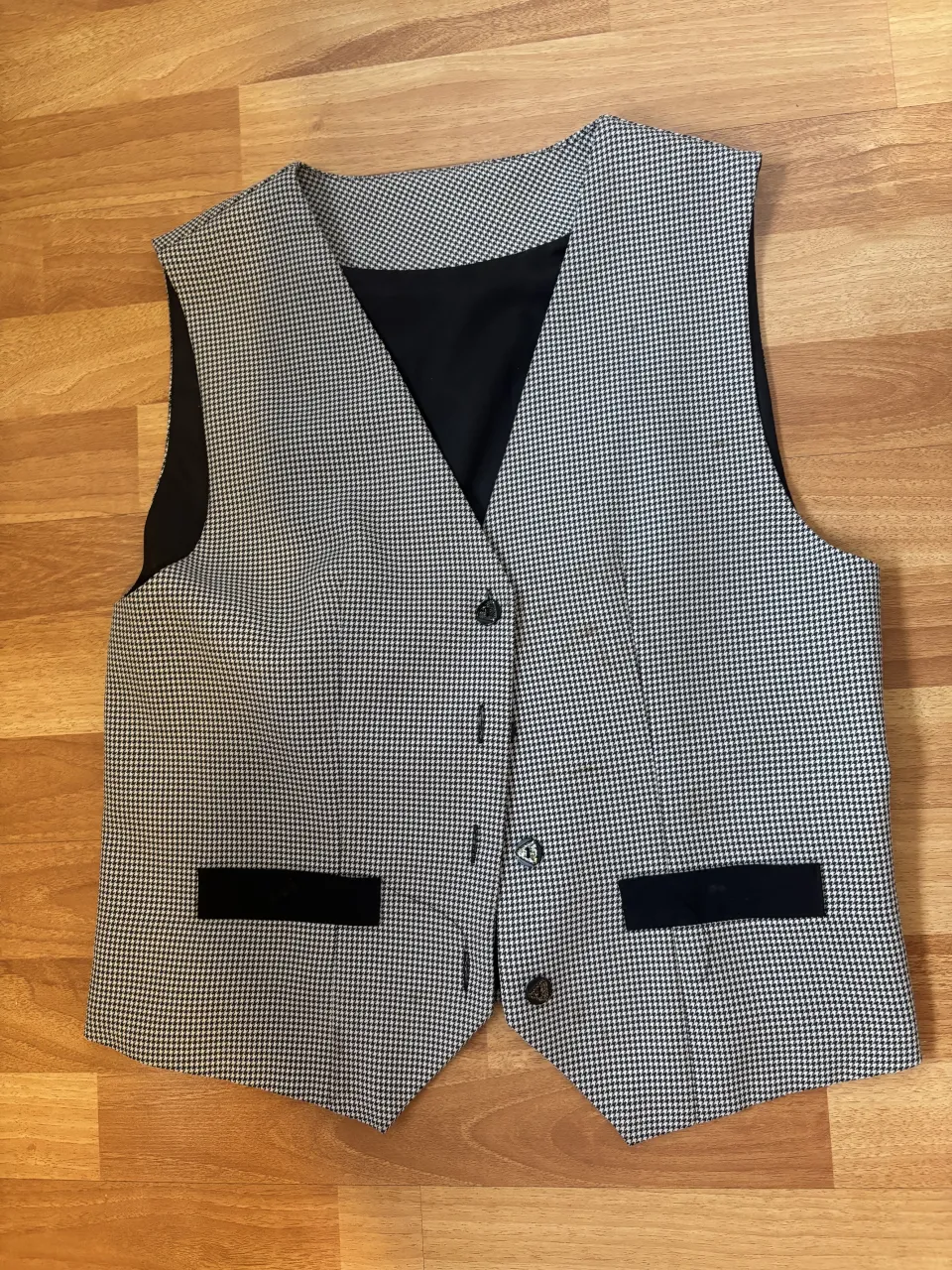Chaleco de vestir a cuadros pequeños en blanco y negro. Posee dos bolsillos falsos en la parte delantera con solapa negra. En la espalda tiene un detalle de ajuste con hebilla circular y detalles de strass.