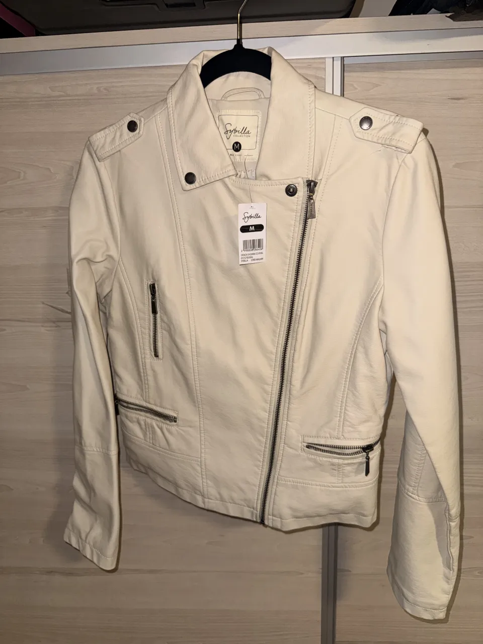 FALABELLA. Campera de cuero sintético color crema, estilo biker. Tiene cuello con solapas, cierre asimétrico al frente, bolsillos con cierre en el frente y en el pecho, y detalles de tachas en los hombros.