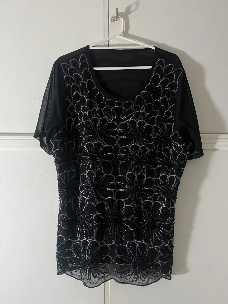 Remera negra con mangas cortas de tul y apliques de flores bordadas y con mostacillas en plateado. Posee cuello redondo y terminación ondulada en la parte inferior.
