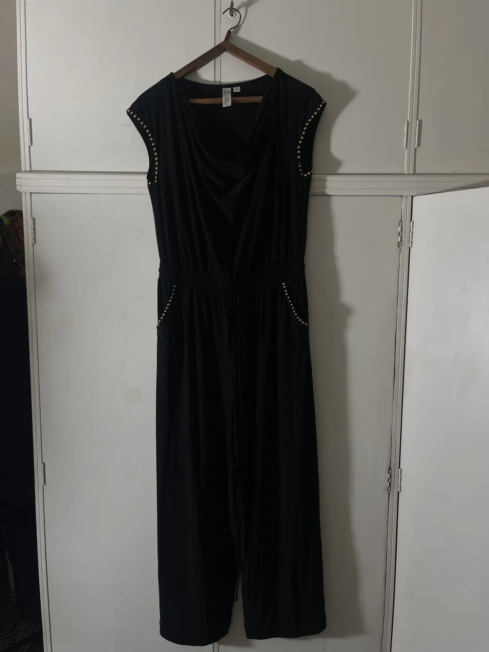 Mono negro con detalles de tachas doradas en los hombros y bolsillos. Posee cuello drapeado y cordón de ajuste en la cintura.