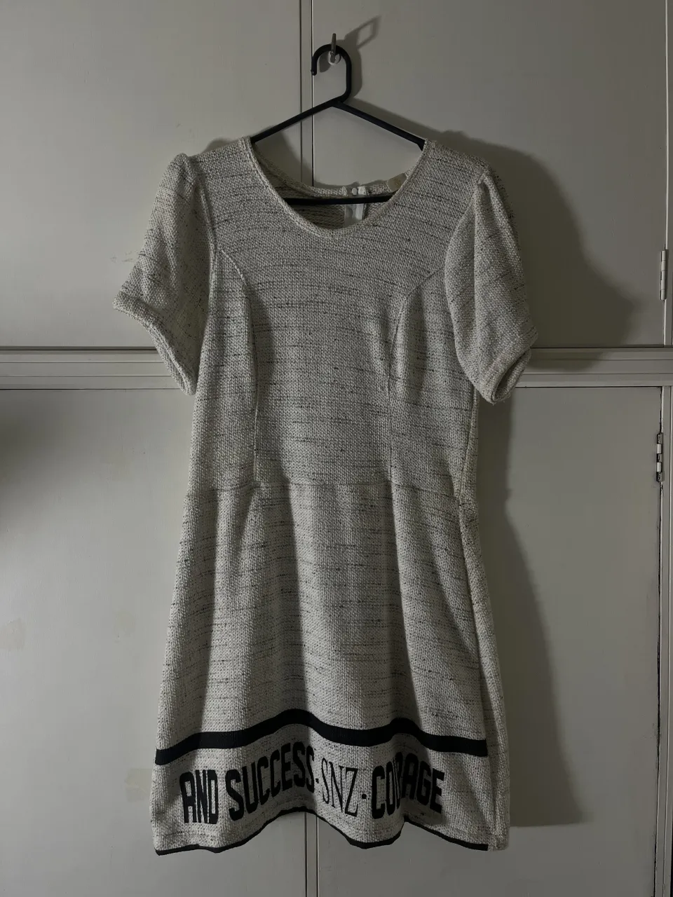 Vestido corto de tejido texturizado blanco y negro con estampa de letras negras en la parte inferior. Posee mangas cortas y cuello redondo.