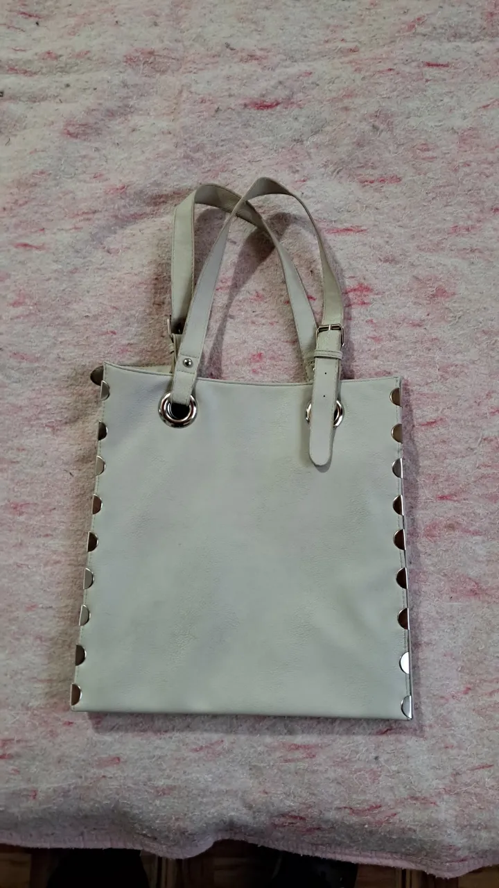 Bolso blanco de cuero con tachas plateadas en los laterales y manijas regulables. Posee múltiples compartimentos internos, uno de ellos con cierre.
