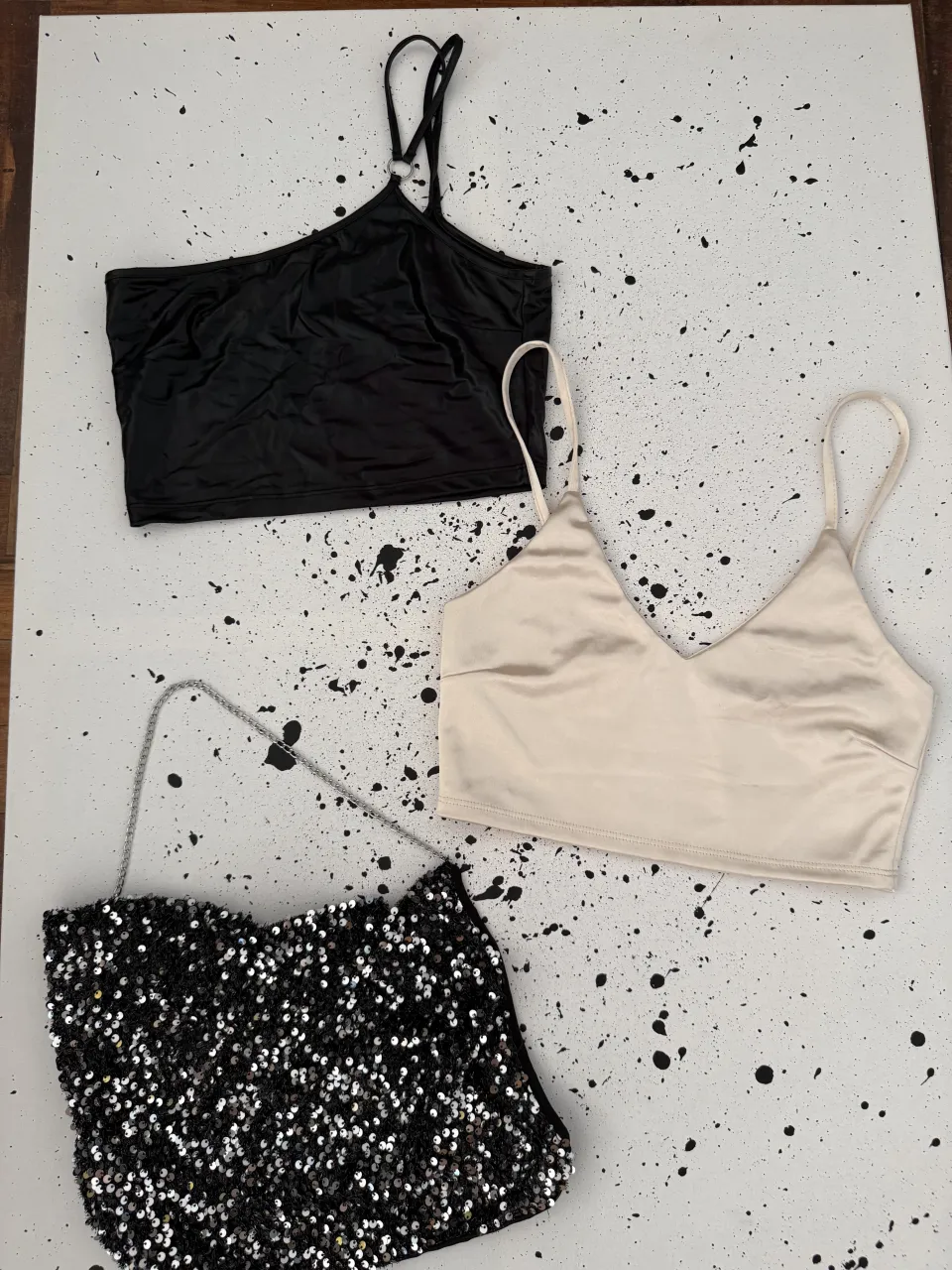 Conjunto de dos tops y una cartera. Un top negro con tiras finas y detalle metálico en el cuello. Un top color crema con escote en V y tiras finas. Cartera negra con lentejuelas plateadas y cadena.