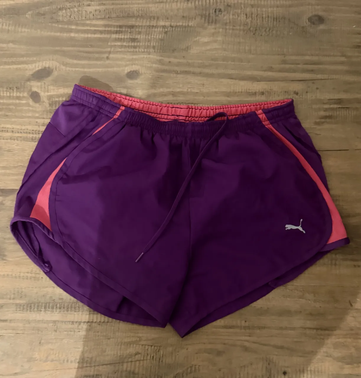 Shorts deportivos de color púrpura con detalles en rosa. Cintura elástica con cordón de ajuste. Logo de Puma en el lateral.