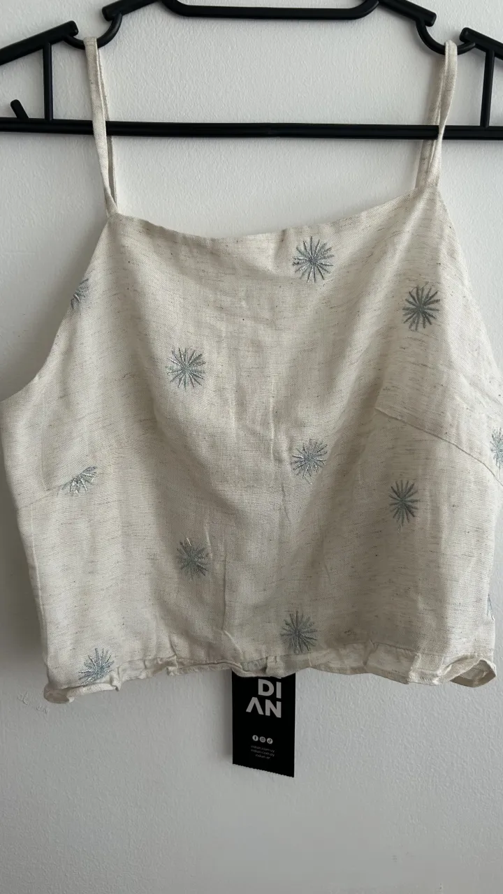 Musculosa de tela color crudo con estampa de flores bordadas en color celeste. Posee breteles finos y espalda con fruncido.