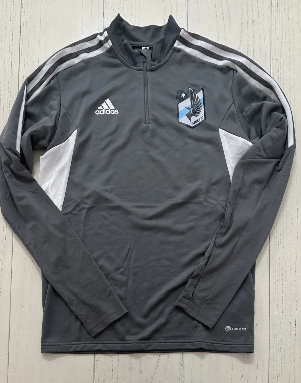 Buzo deportivo gris de manga larga con cierre hasta el cuello y logo de Adidas y equipo de fútbol estampado en el frente. Posee detalles blancos y las tres tiras icónicas de la marca en las mangas. La tecnología Aeroready indica que es transpirable.