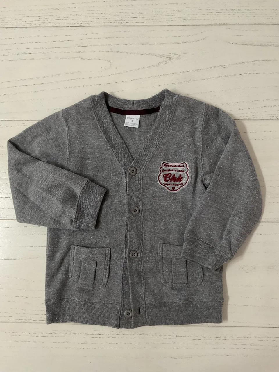 Cardigan gris de algodón con cuello V, botones al frente y dos bolsillos tipo parche. Tiene un escudo bordado en el pecho izquierdo con la inscripción "Cheeky Original Clothing". Ideal para un look casual y cómodo.
