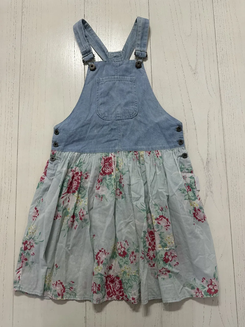 Vestido de jean con tiradores y falda estampada con flores rosas y verdes sobre fondo celeste. Tiene dos bolsillos en el frente y botones a los costados.