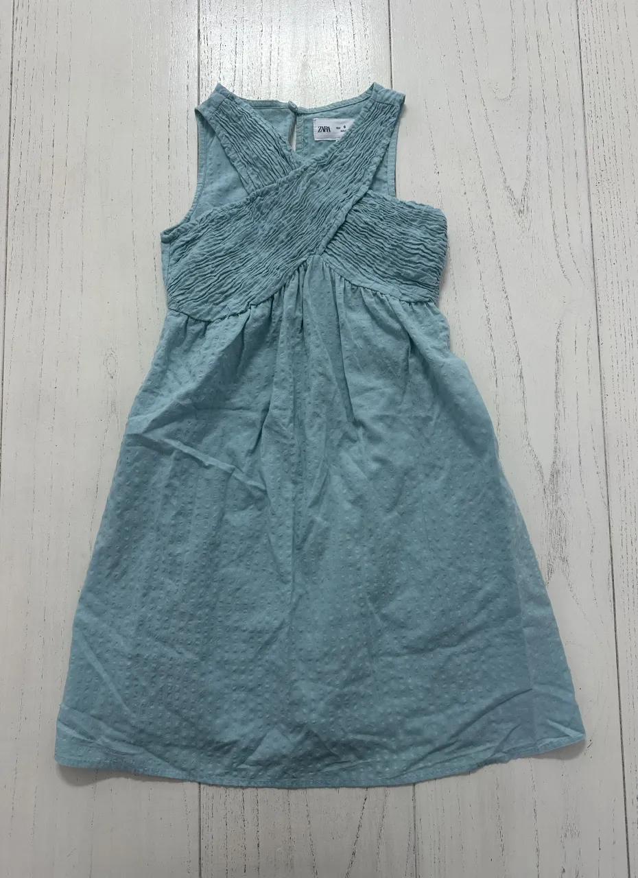 Vestido celeste con textura de pequeños puntos y detalles fruncidos en la parte superior. Posee tiras cruzadas en el escote y un botón en la espalda.