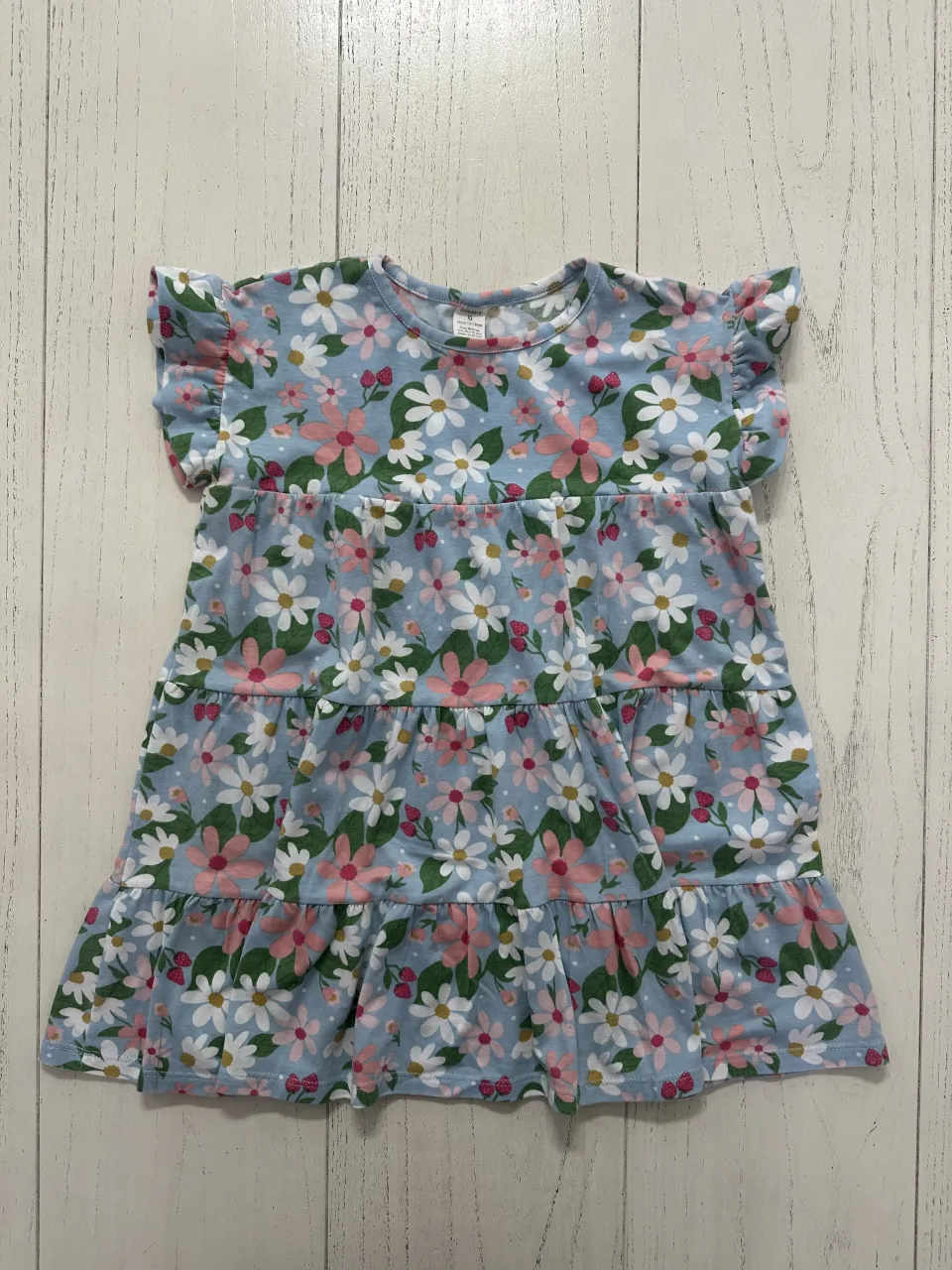 Vestido celeste con estampa de flores blancas, rosas y detalles en rojo. Posee volados en las mangas y tres frunces horizontales en la falda.