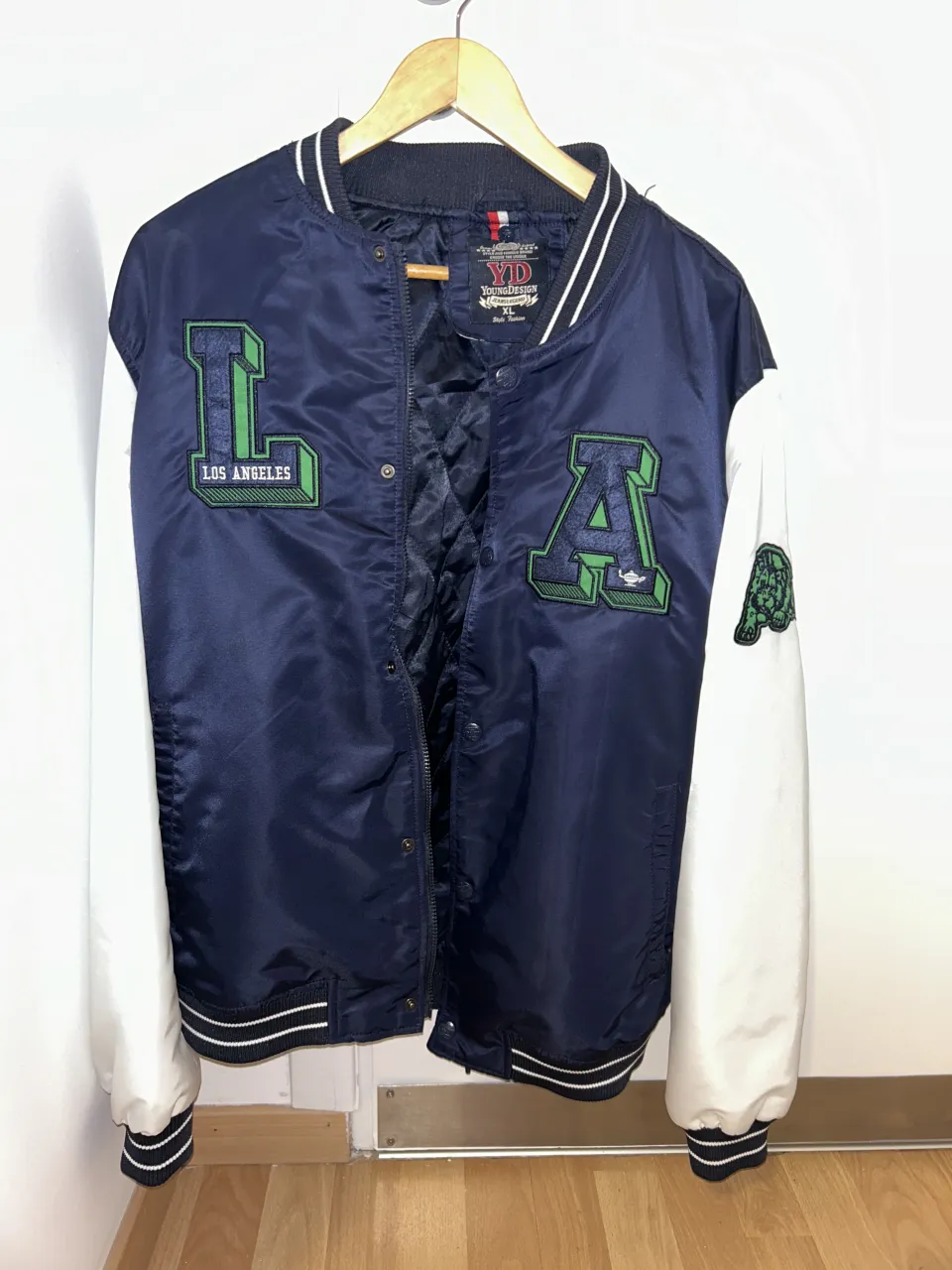 Campera bomber de estilo universitario, con mangas blancas y cuerpo azul marino. Presenta parches bordados de 'LA' y un león en la manga. Ideal para un look urbano y canchero.