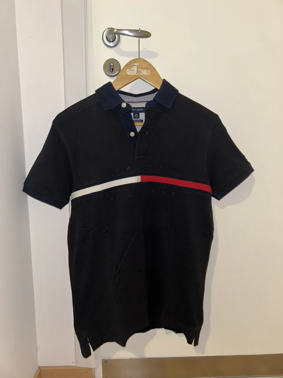 Remera polo negra de manga corta, con detalles de franjas blancas y rojas en el pecho. Logo de la marca visible. Ideal para un look casual y moderno.