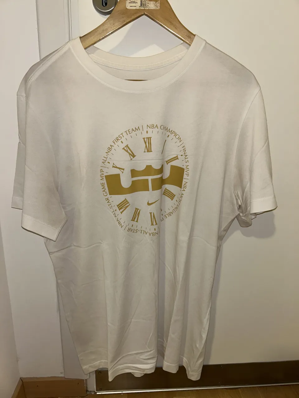 Remera blanca de manga corta con un estampado dorado en el frente que simula un reloj con números romanos y texto relacionado a logros de básquet. Ideal para un look casual y deportivo. Las imperfecciones que se le ven son por culpa de la luz, la prenda está en muy buen estado.