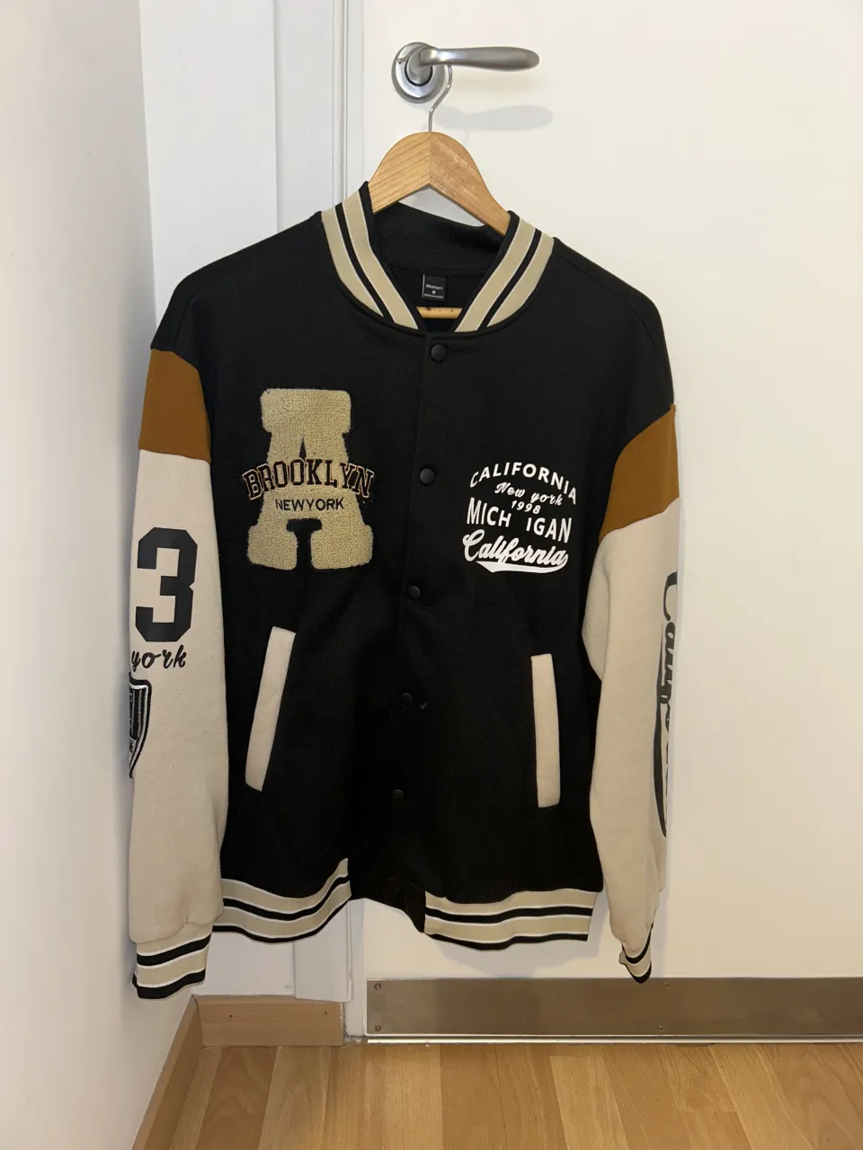 Campera estilo universitaria, negra con mangas color crudo y detalles en marrón. Tiene bordados y estampas de ciudades como "Brooklyn", "New York", "Michigan" y "California". Ideal para un look casual y urbano.