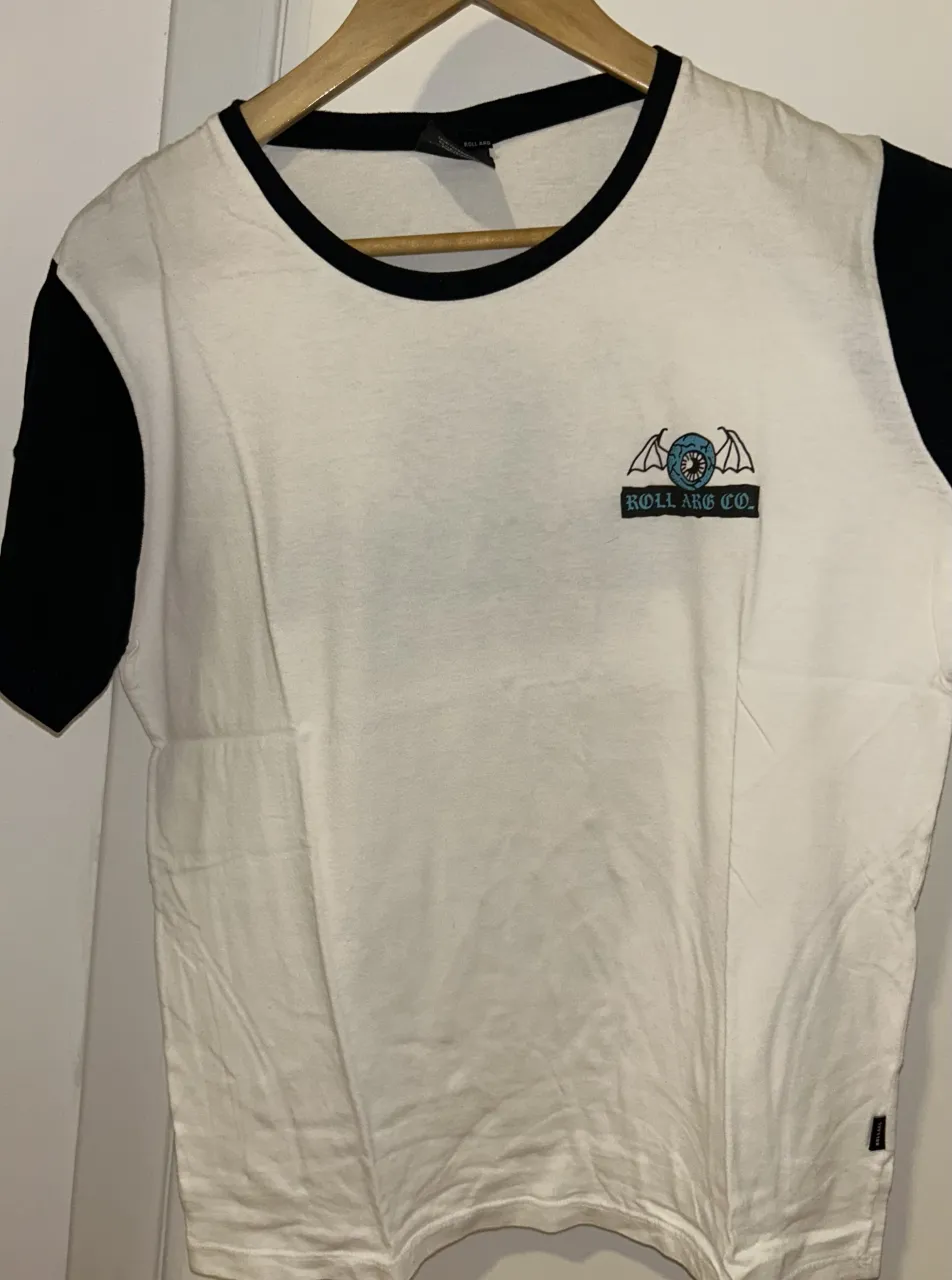 Remera blanca con mangas negras y estampa de ojo con alas y logo "ROLL ARG CO. SKATES" en la espalda. Ideal para un look urbano y casual.