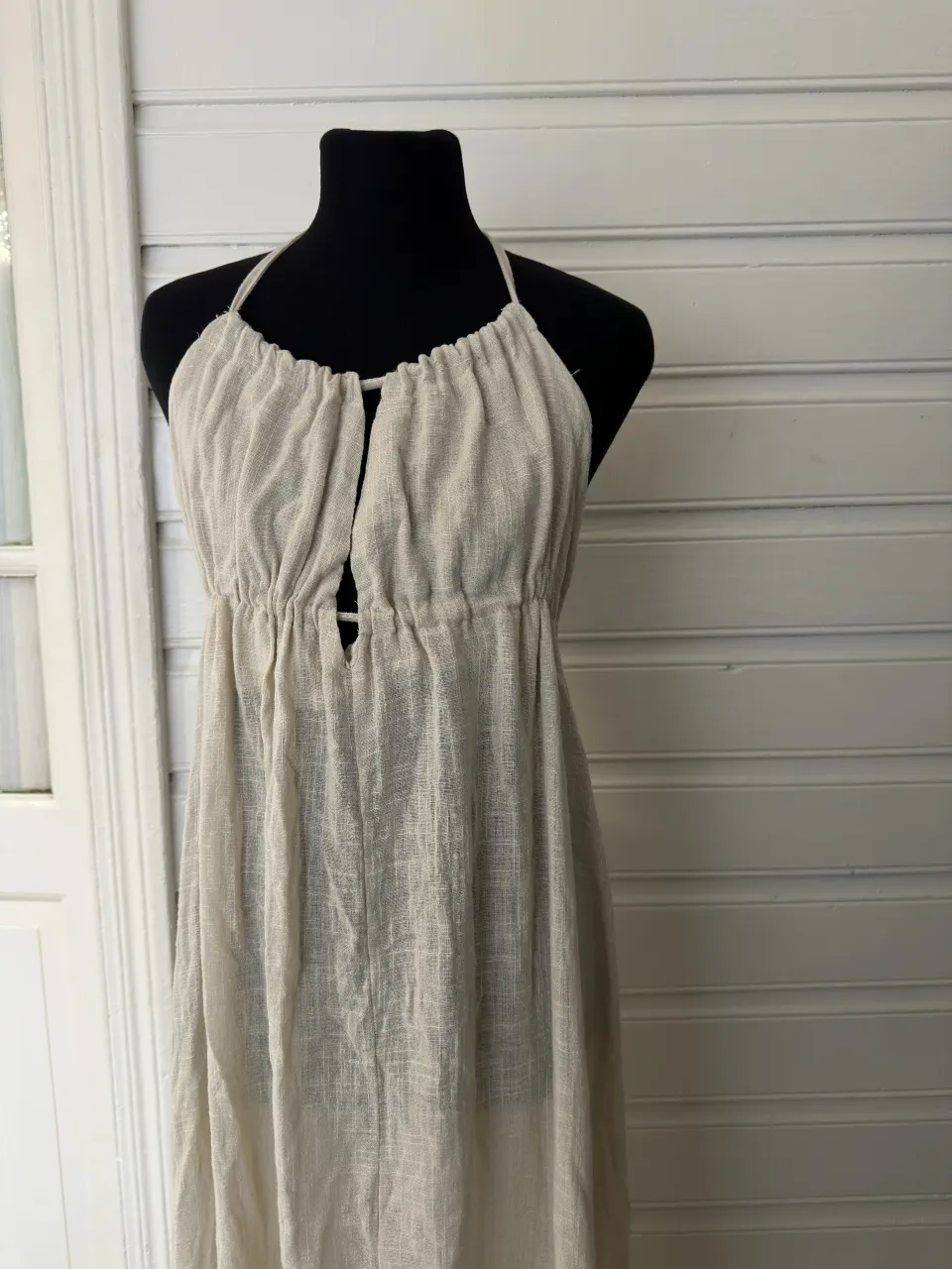 Vestido largo de tela liviana, color crudo. Posee breteles finos que se anudan en la nuca y un recorte en el escote. La parte superior del busto está fruncida y la cintura también.