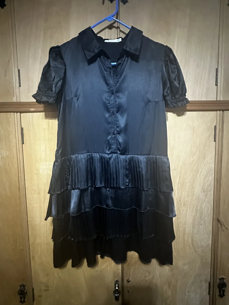 Vestido negro con cuello de camisa y botones al frente. Posee mangas cortas abullonadas y falda con volados y plisados.