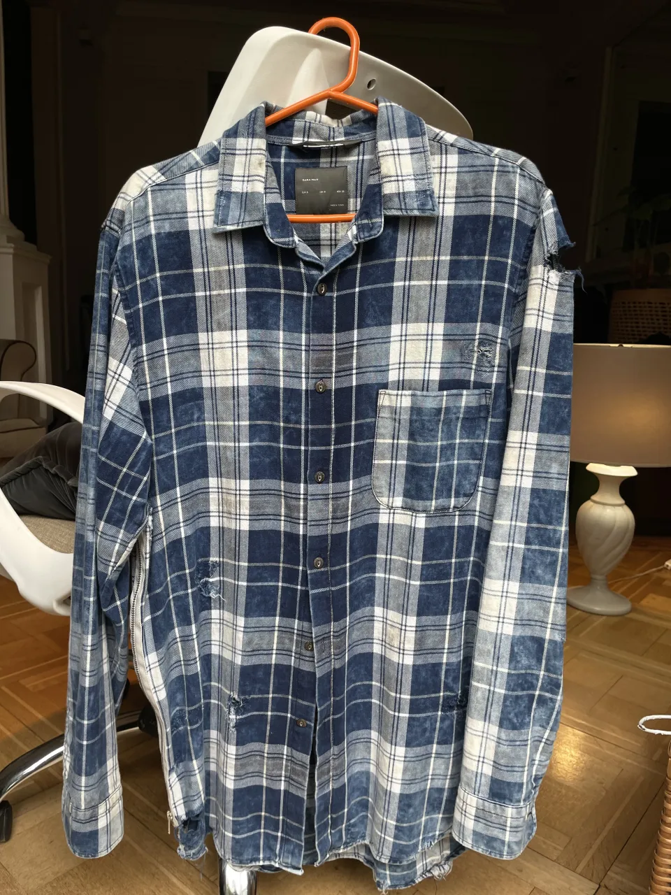 Camisa a cuadros ZARA de manga larga con roturas y detalles de cierre en los laterales. Presenta un estampado de cuadros en tonos azules y blancos, con un diseño desgastado y texto estampado en la espalda.