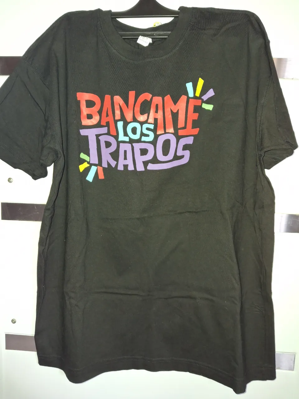 Remera negra de manga corta con estampa "BANCAME LOS TRAPOS" en la parte delantera. La estampa tiene letras en rojo, violeta y celeste con detalles de colores vibrantes.