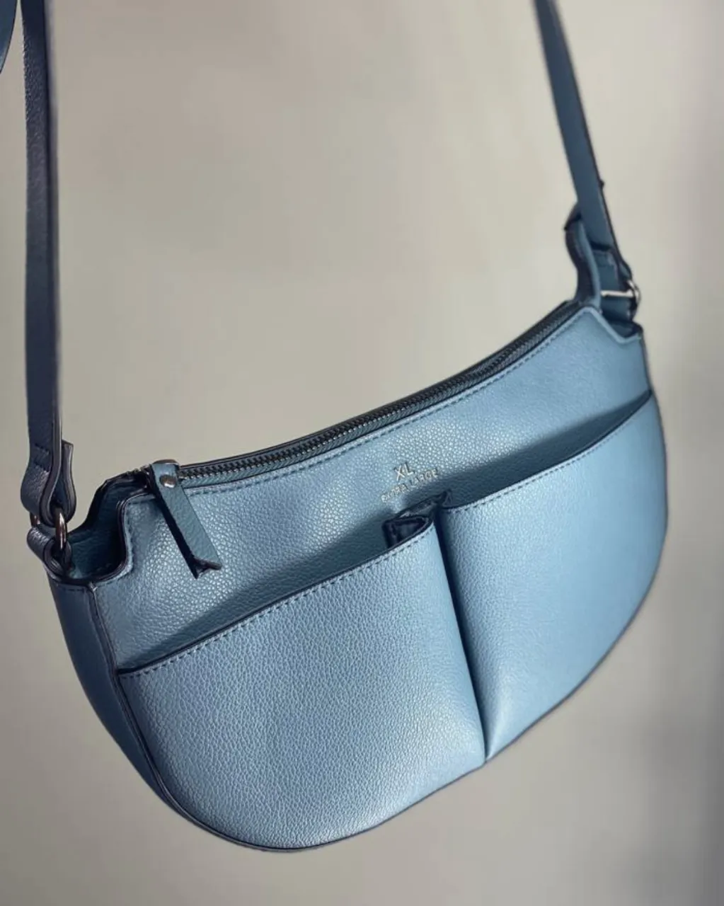 Cartera marca XL nueva sin uso. color azul claro con correa de hombro. Tiene un bolsillo exterior con cierre en la parte trasera y dos bolsillos exteriores abiertos en la parte delantera. '.