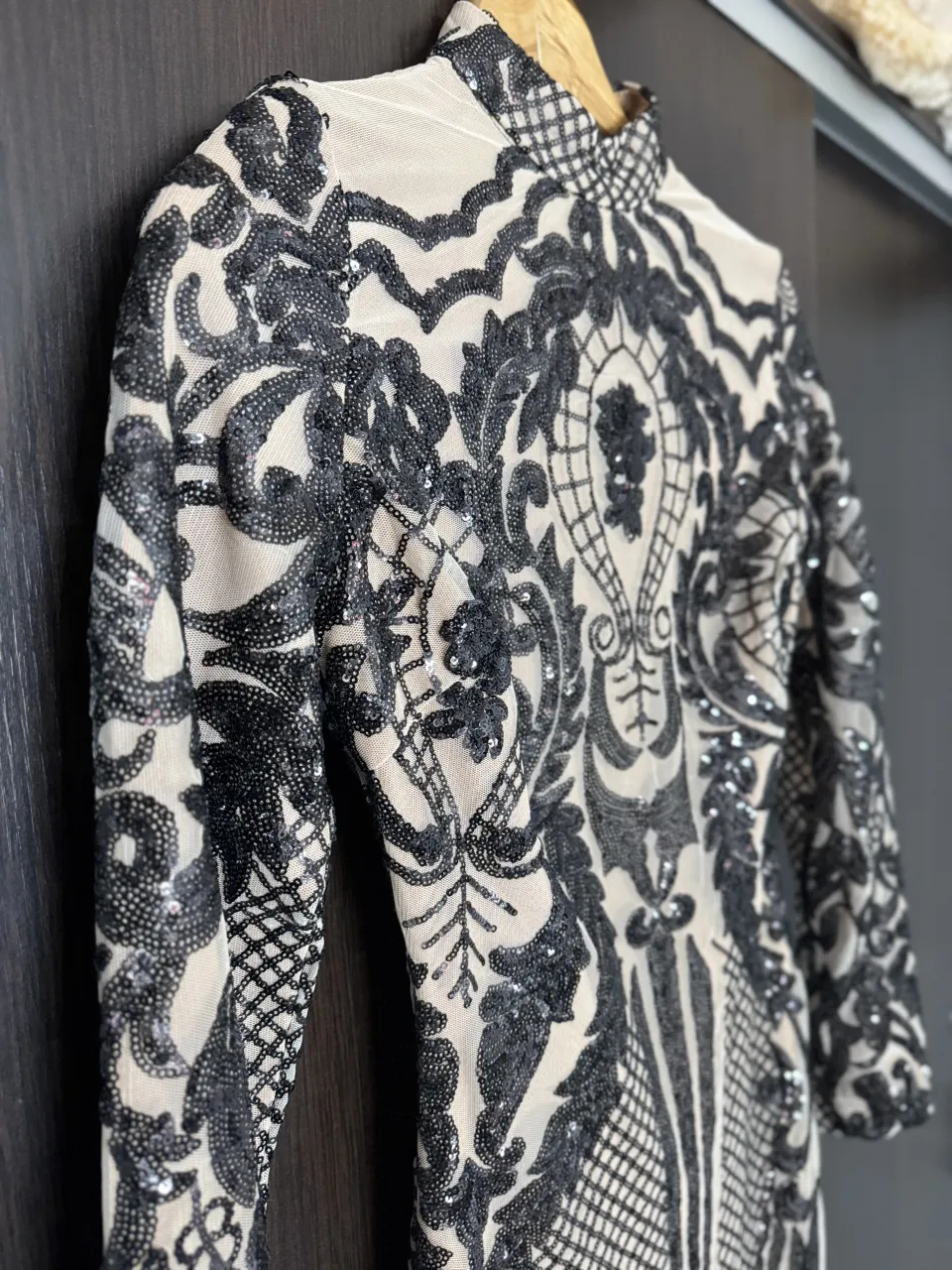 Vestido corto con cuello alto y mangas largas. Posee un estampado floral y geométrico en negro sobre una base color piel. El diseño está cubierto de lentejuelas.