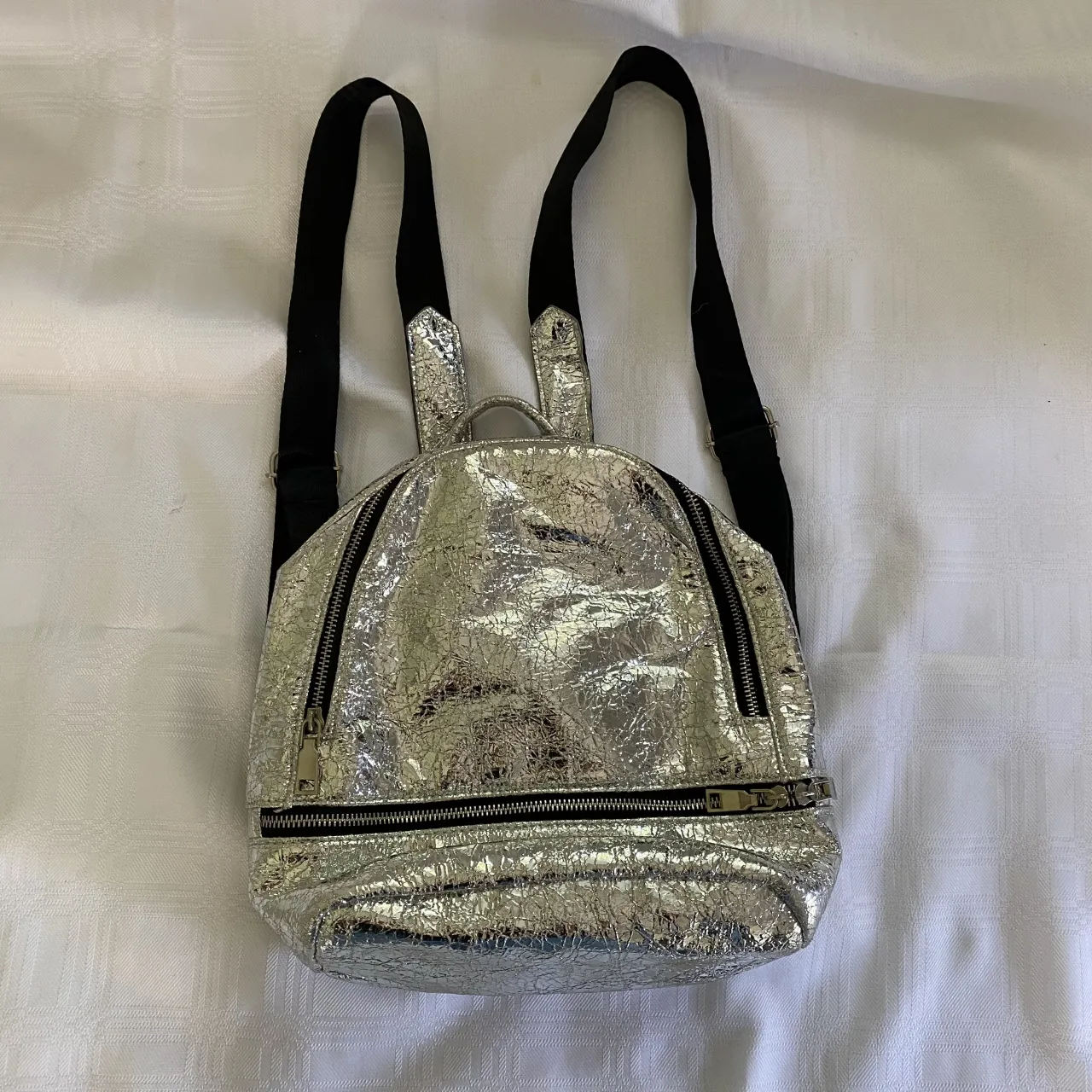 Mochila plateada con textura arrugada y dos cierres frontales. Las correas son negras.